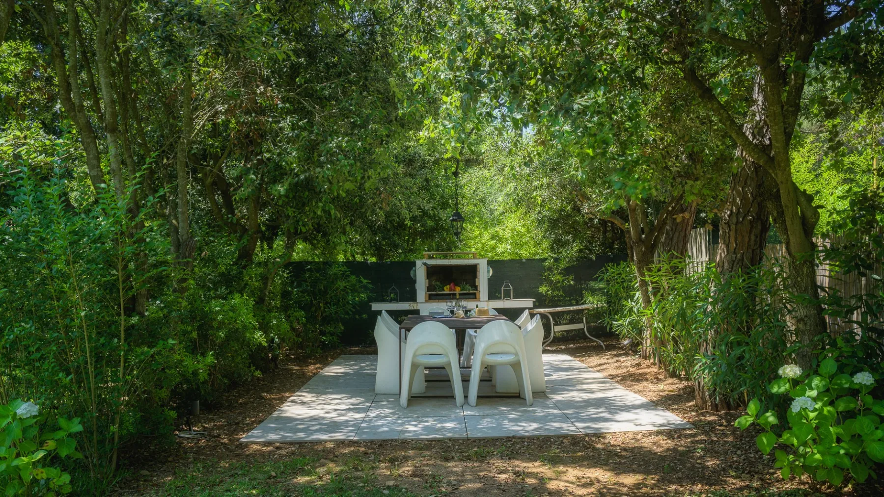 A dining table and chairs sit on a paved patio beneath leafy trees.