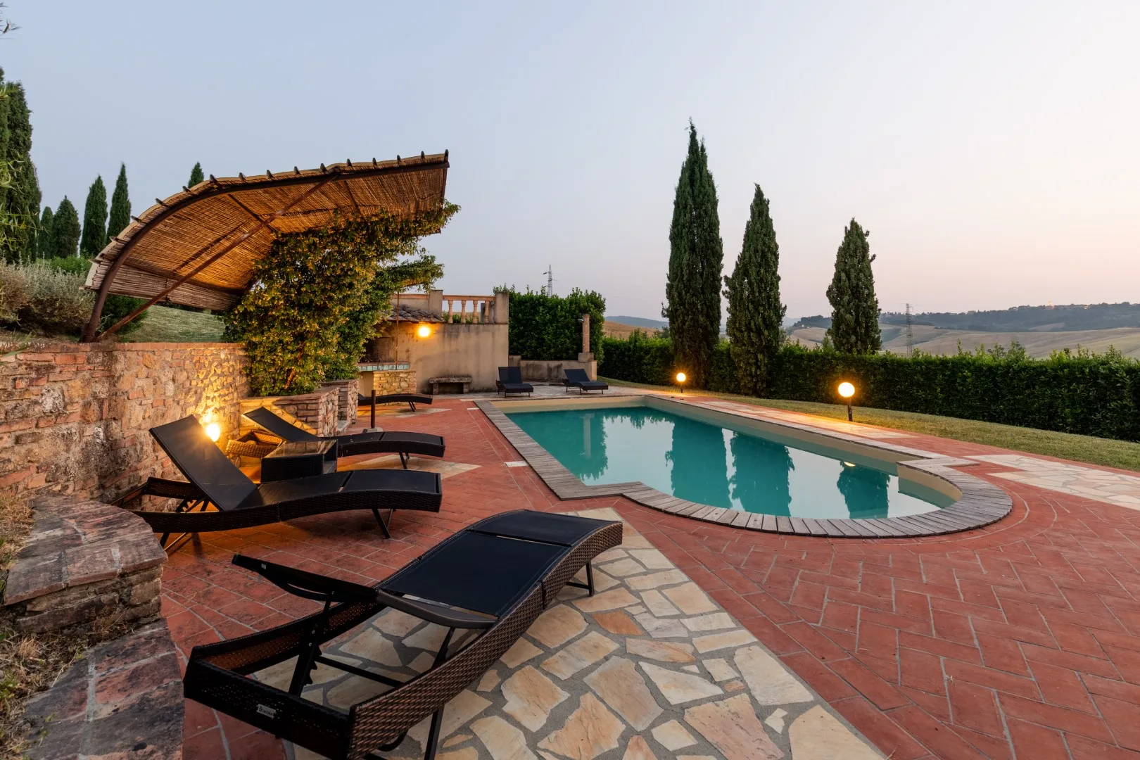 An outdoor swimming pool beside a paved patio with lounge seating and trees.