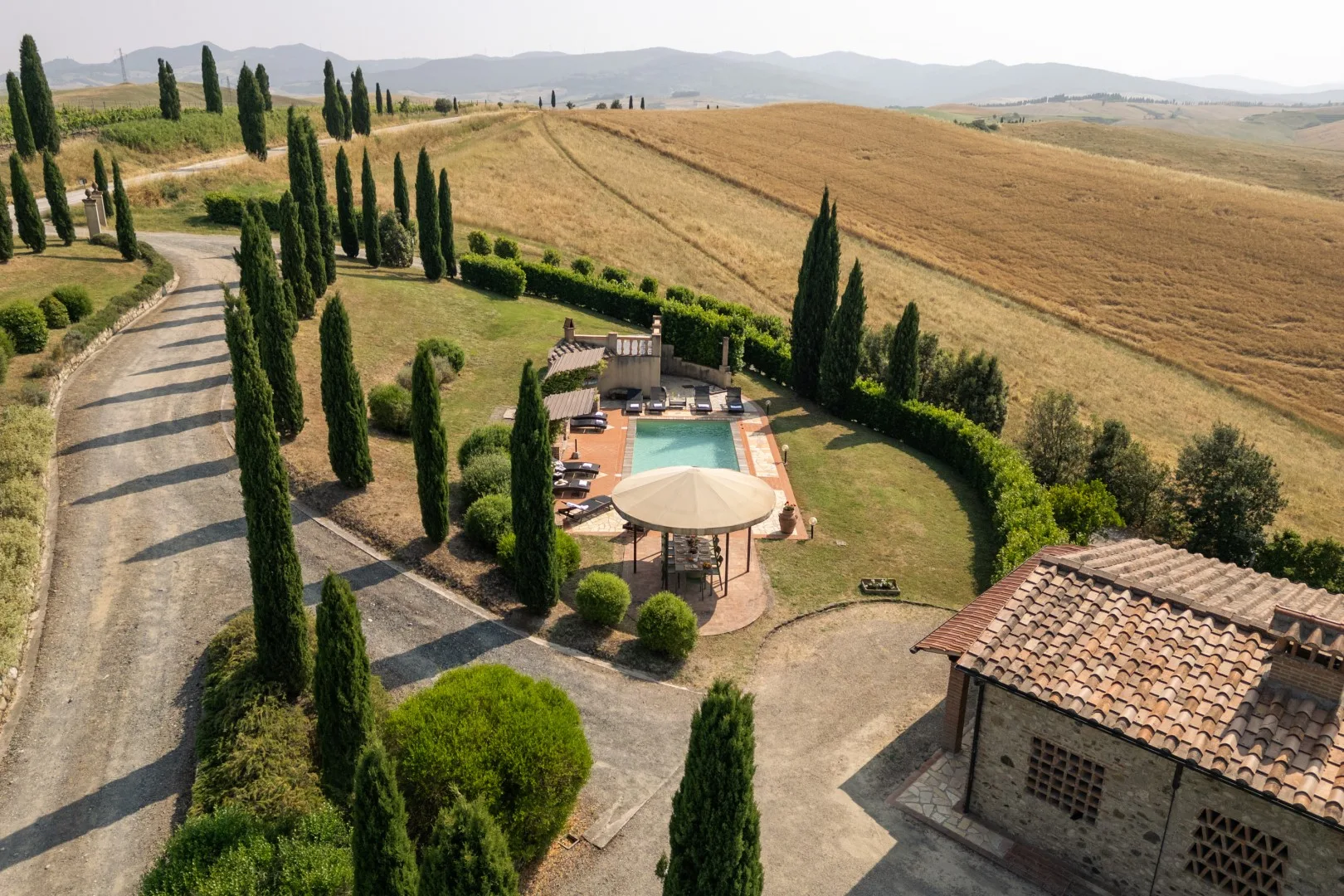 A countryside pool area with cypress trees, beside a tiled-roof building.