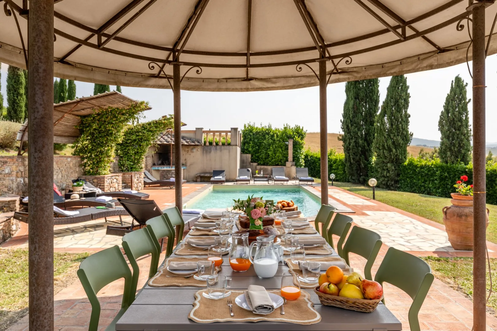 A covered outdoor dining table overlooks a swimming pool and garden.