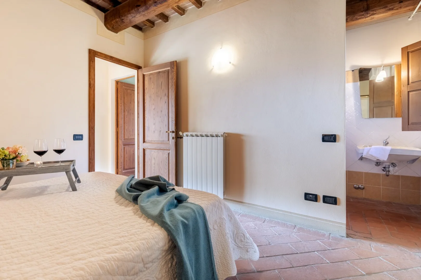 A bedroom with a bed, wooden ceiling beams, and an open bathroom area.