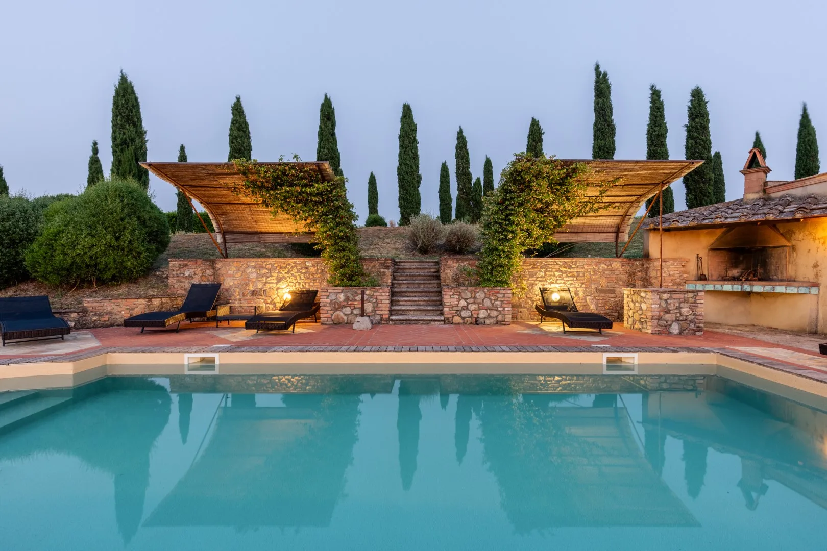 An outdoor swimming pool faces stone walls, greenery, and shaded seating areas.