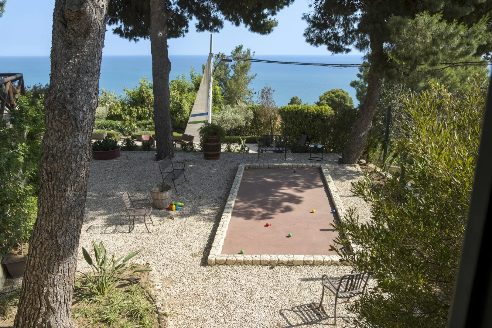 Un jardín de grava con una zona de juego rectangular, árboles y el mar al fondo.