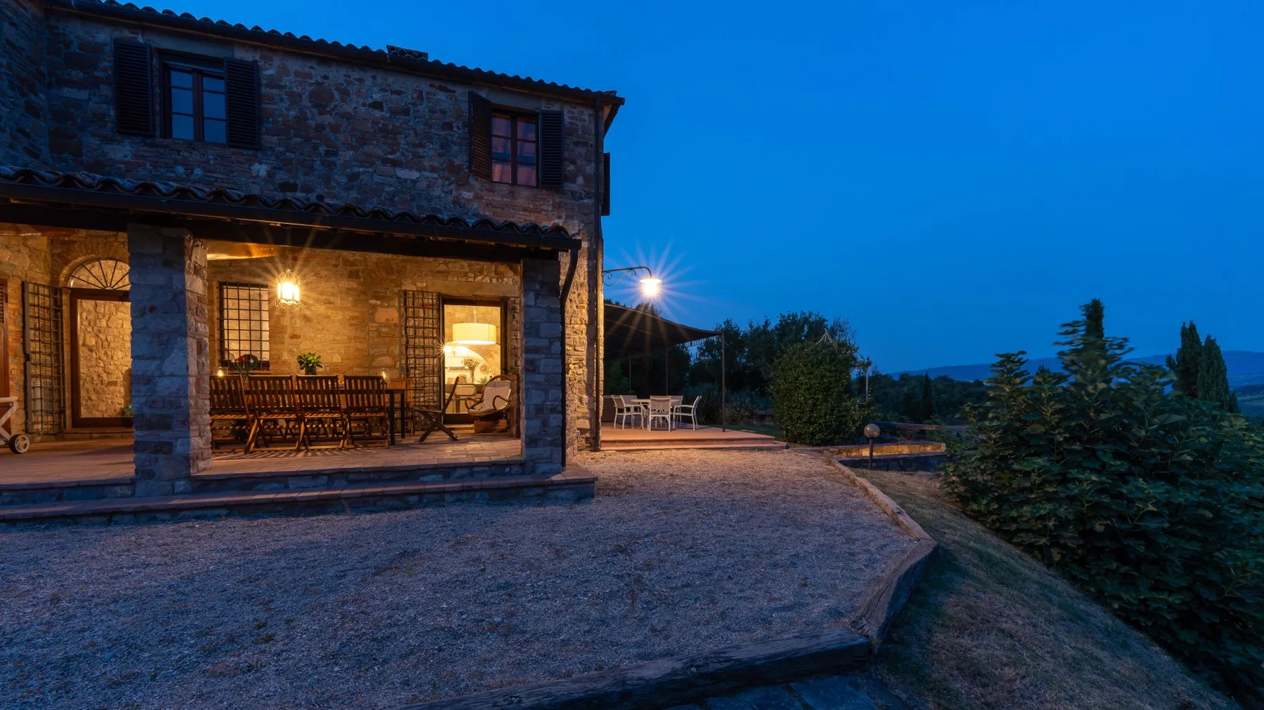 A stone house opens onto a broad paved terrace at dusk.