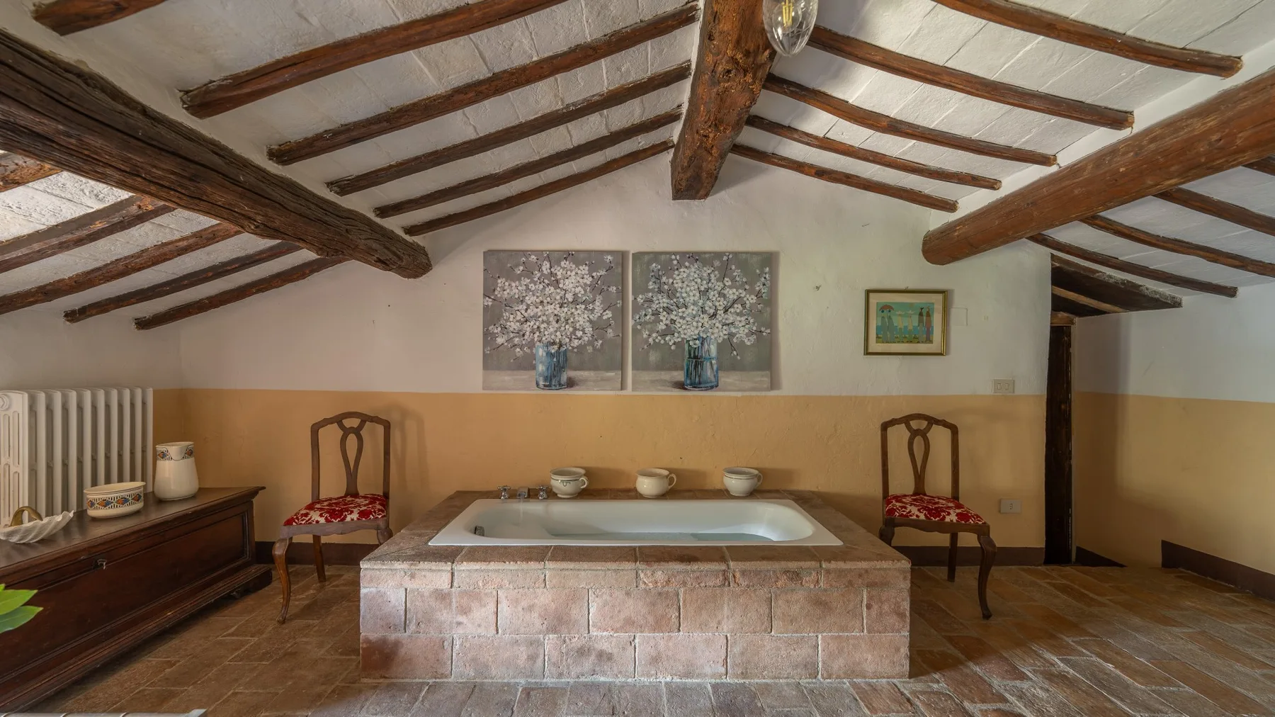 A rustic bathroom features a large tiled bathtub beneath exposed wooden ceiling beams.