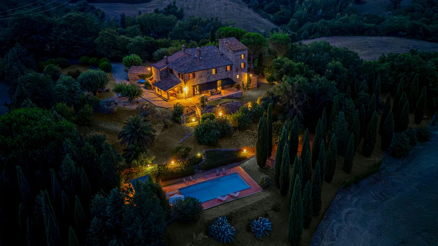 A warmly lit countryside house sits among trees above an outdoor swimming pool.
