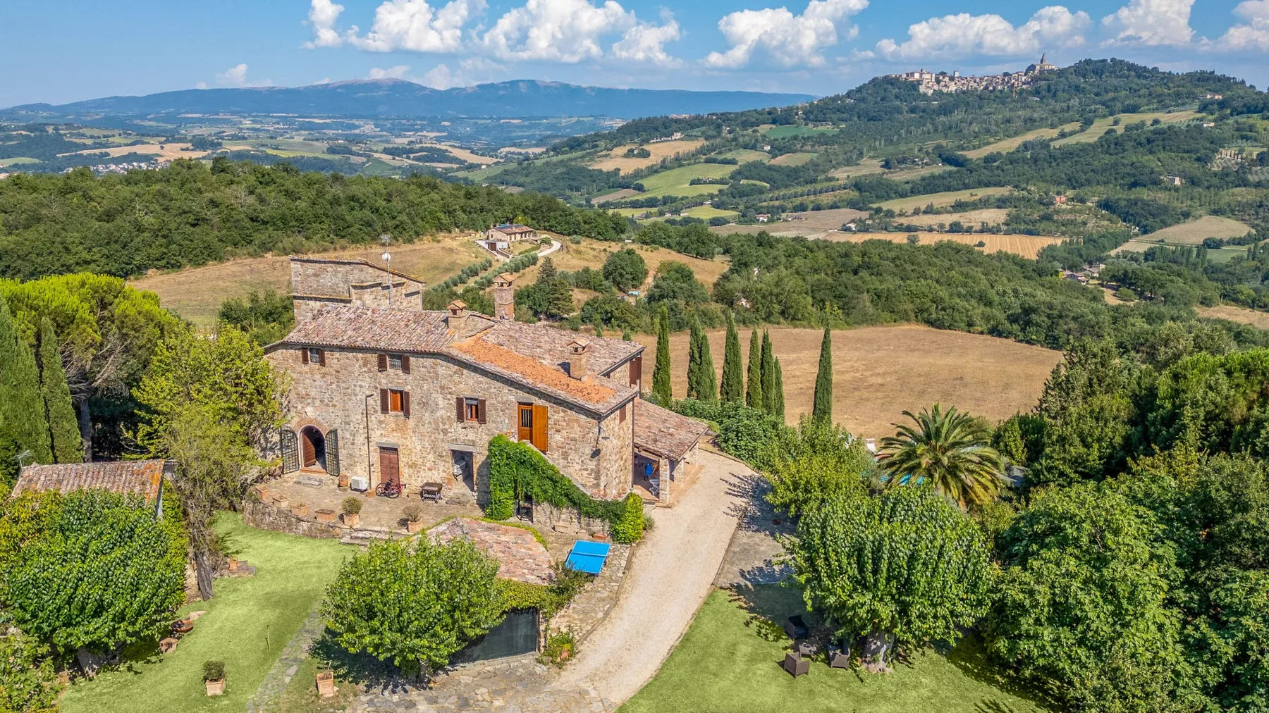 A stone farmhouse sits among gardens and rolling countryside.