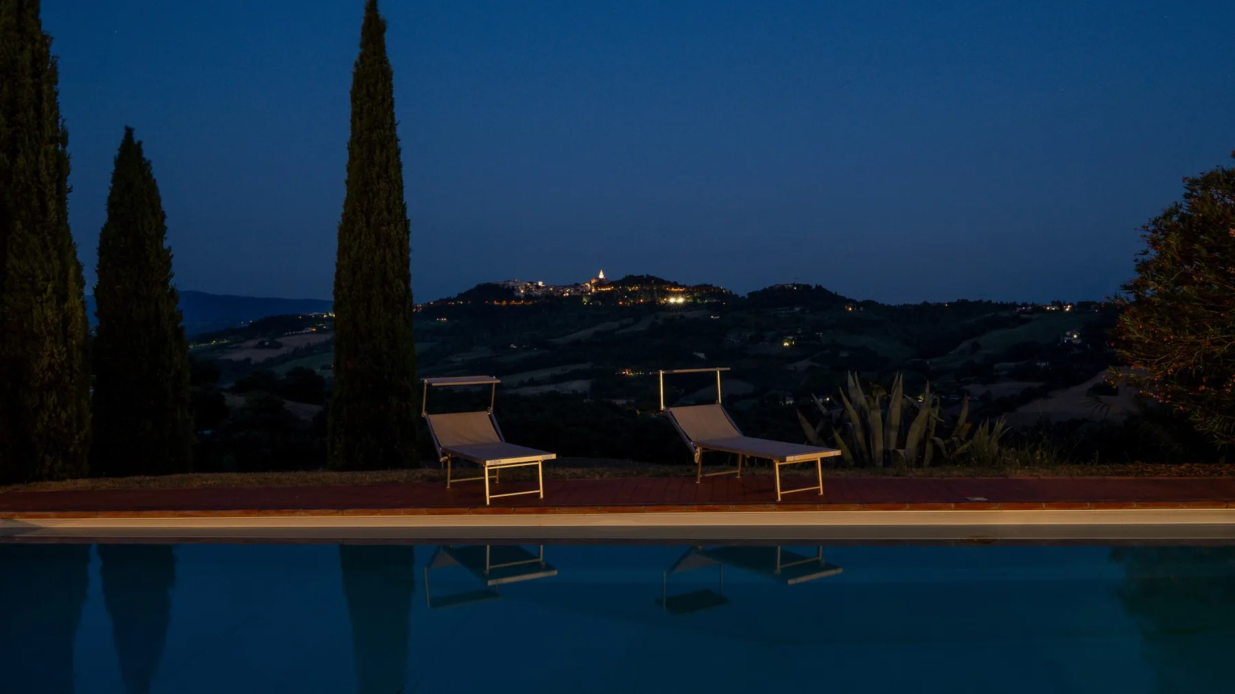 An outdoor swimming pool overlooks dark hills at dusk, with lounge seating nearby.