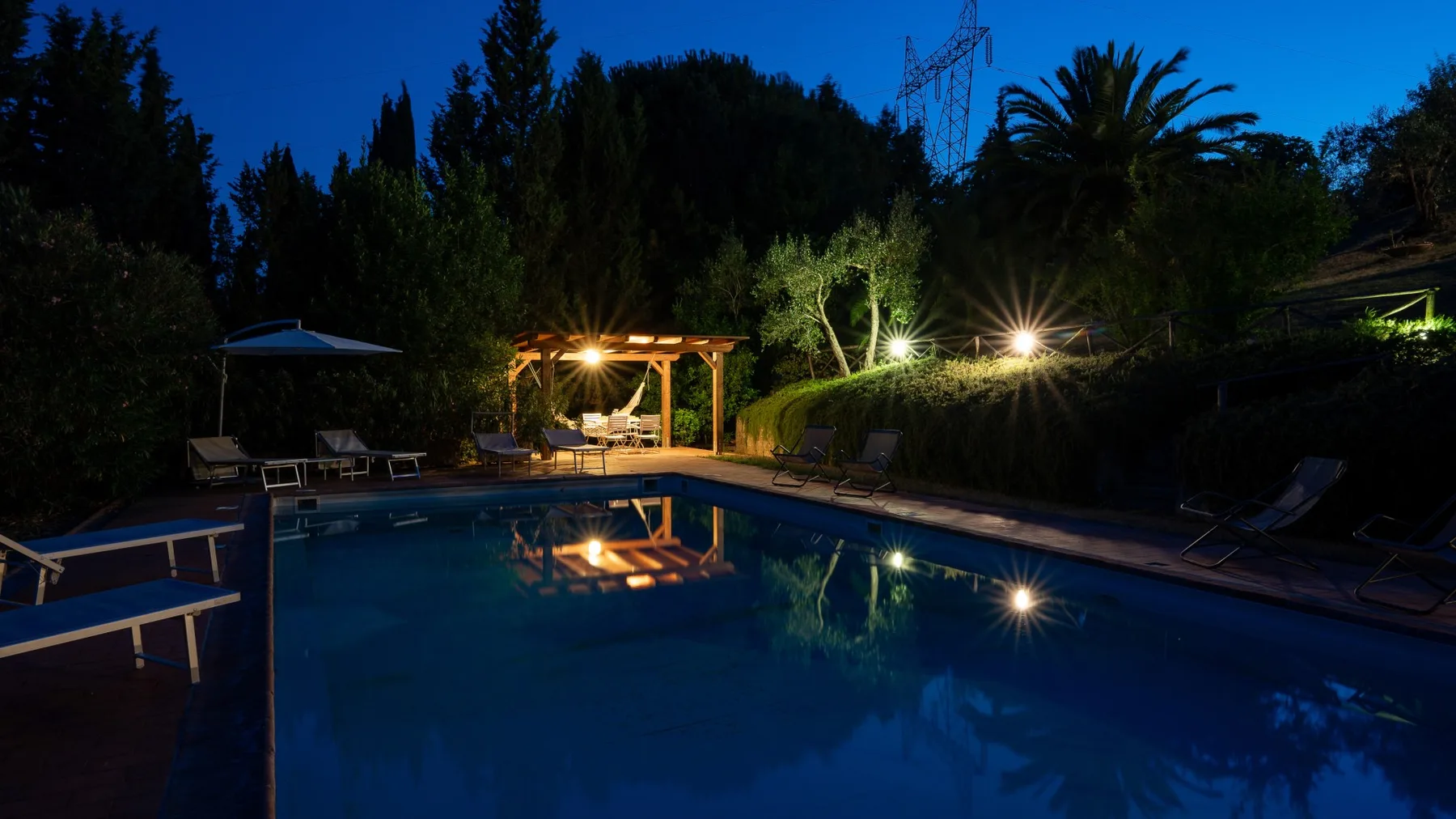 An outdoor swimming pool glows at night, surrounded by trees and lounge seating.