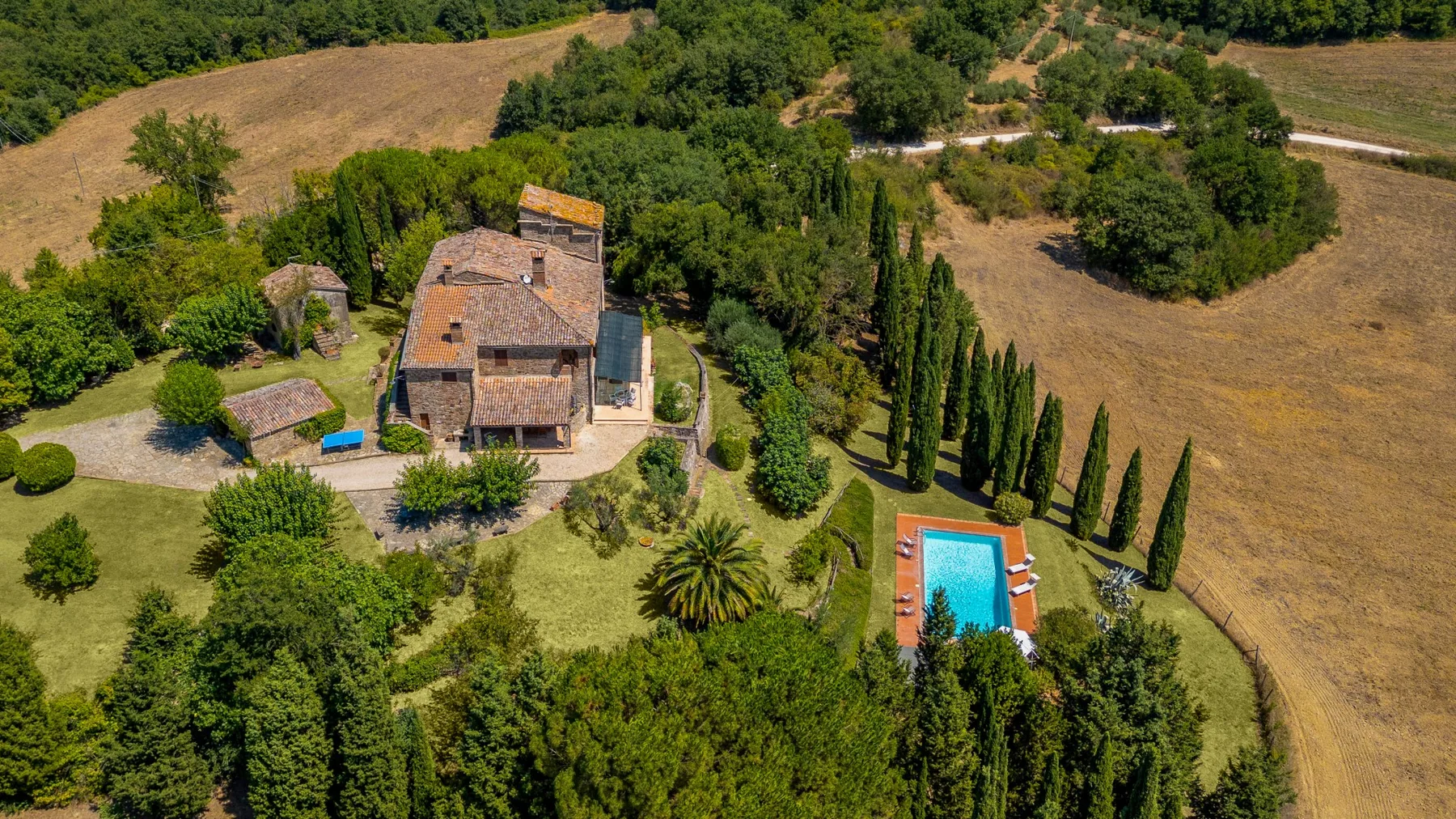 An aerial view of a countryside property with a swimming pool amid trees and open fields.