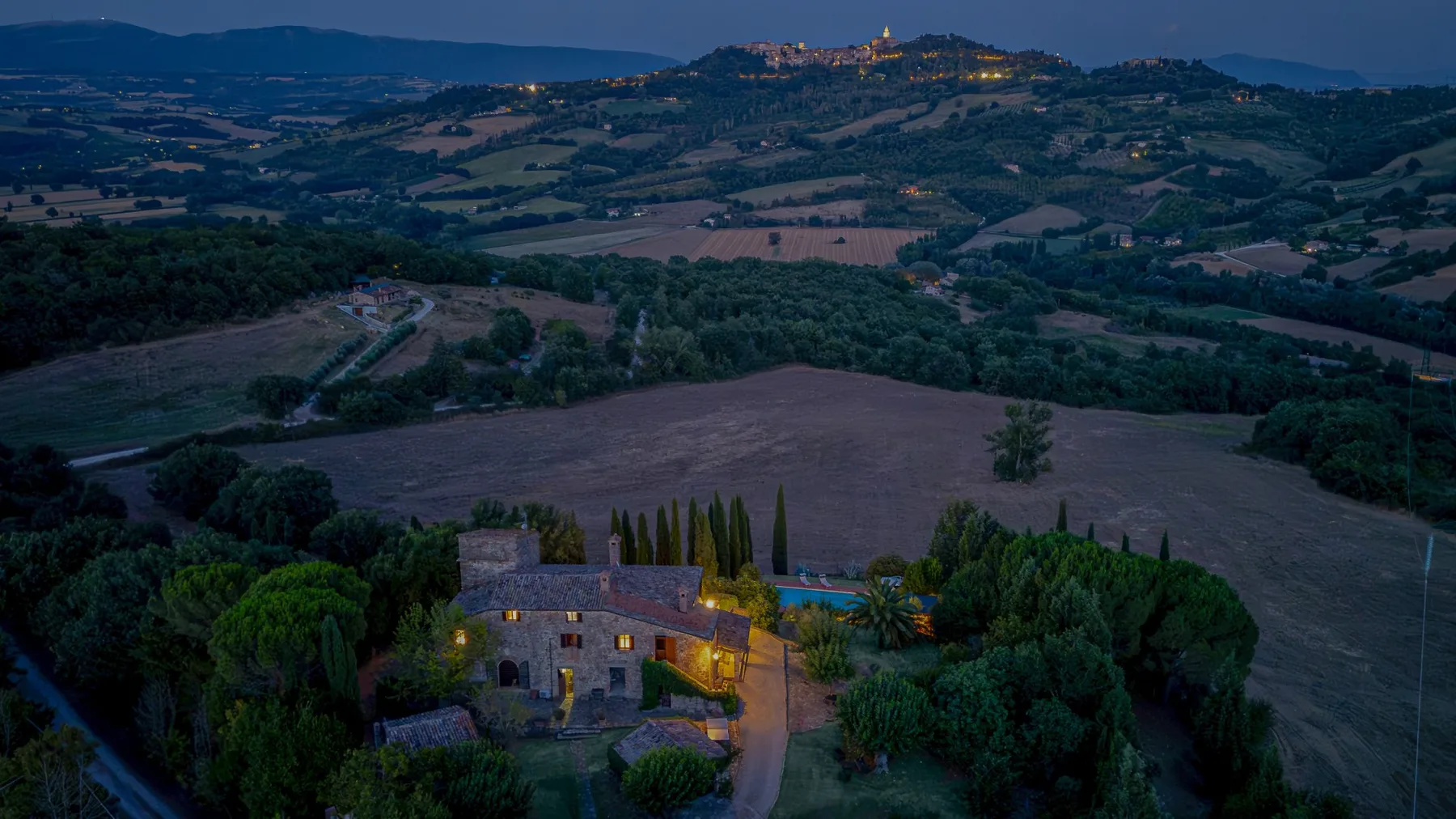A rural landscape at dusk, with a lit stone building among trees and fields.