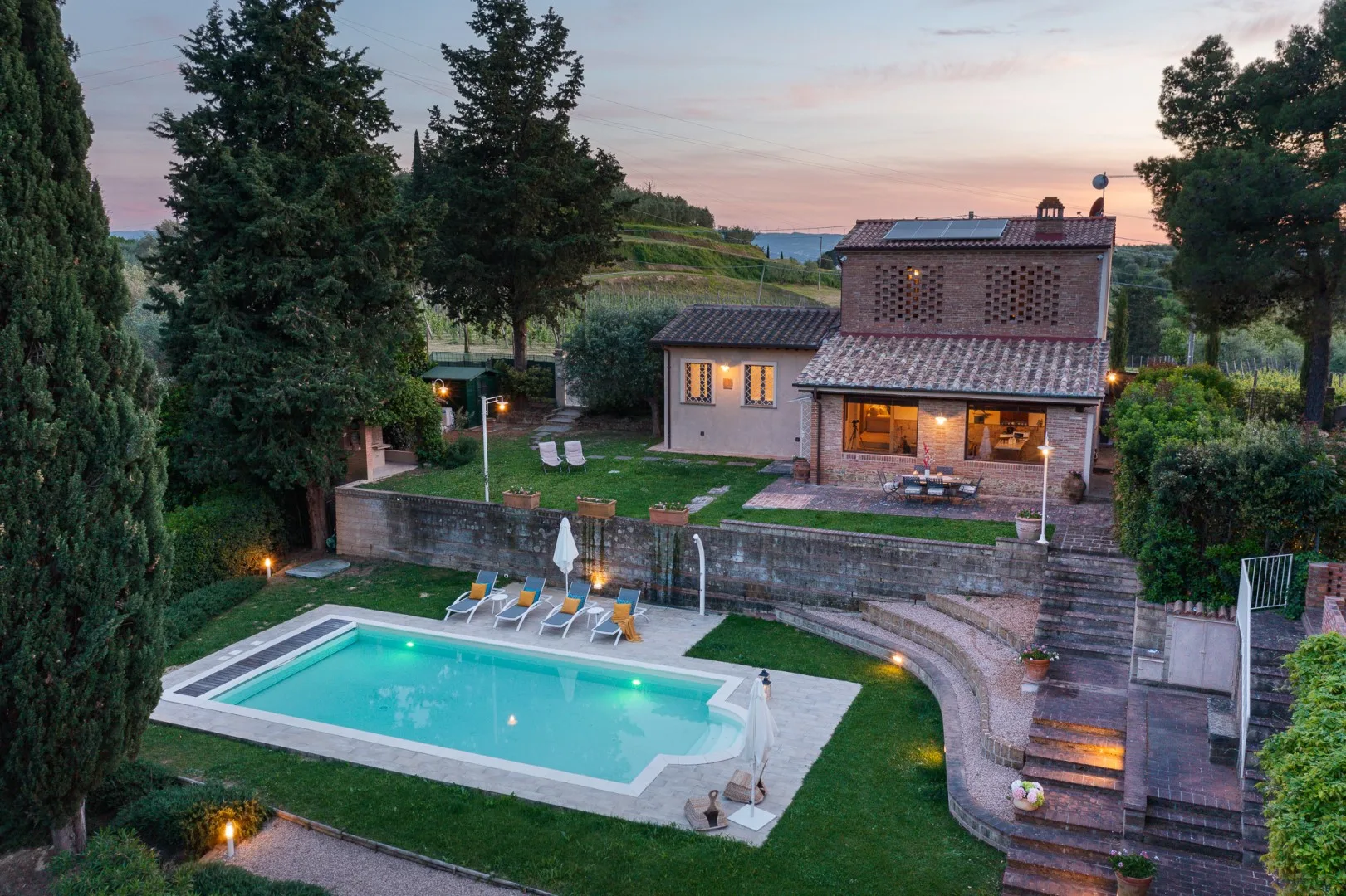 Una casa de campo da a una piscina, un jardín y árboles al anochecer.