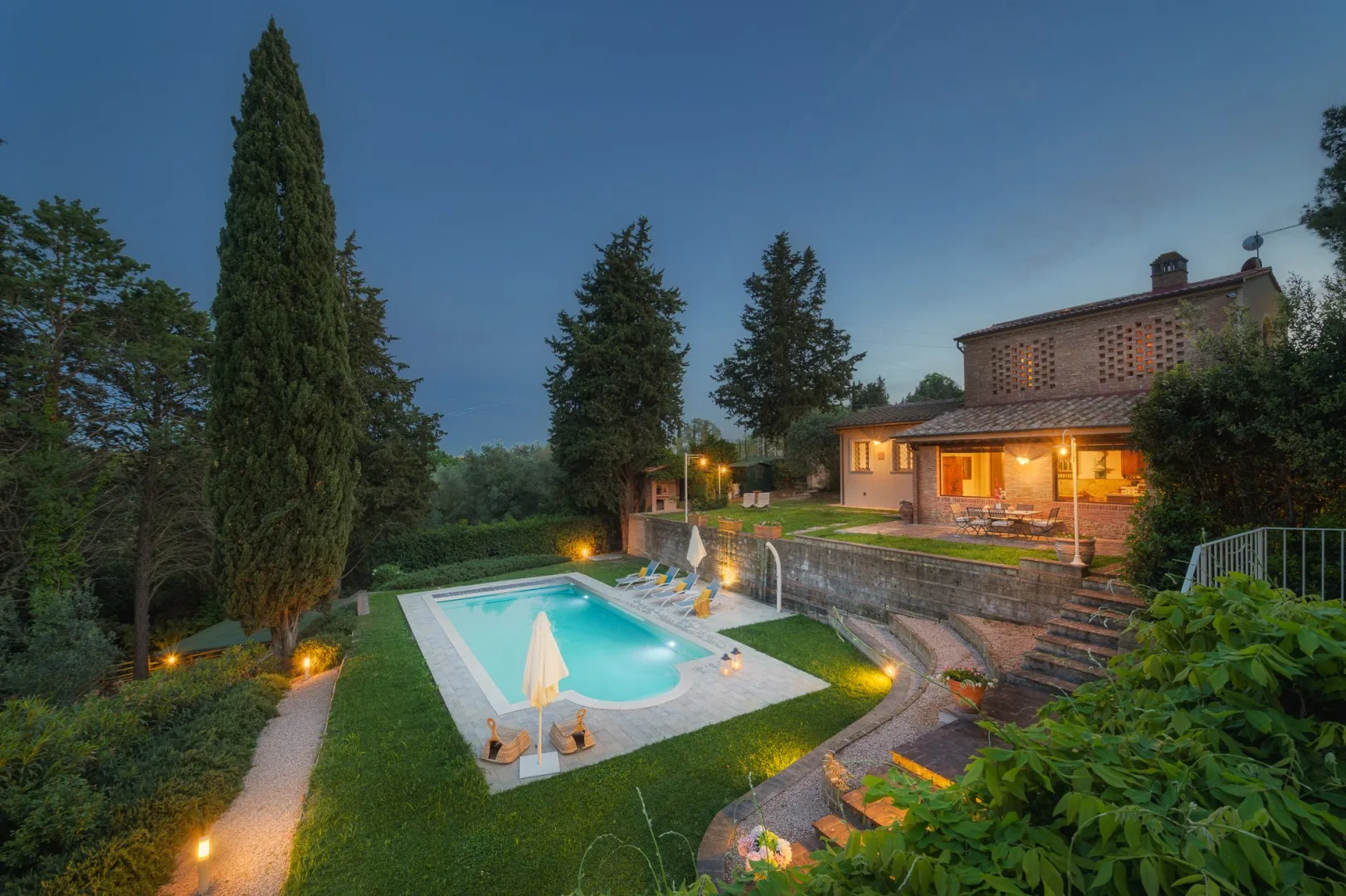 Una piscina al aire libre junto a una casa iluminada y un jardín cuidado.
