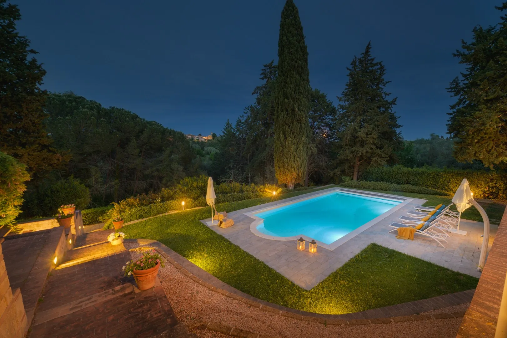 Una piscina iluminada queda junto a un jardín cuidado al anochecer.