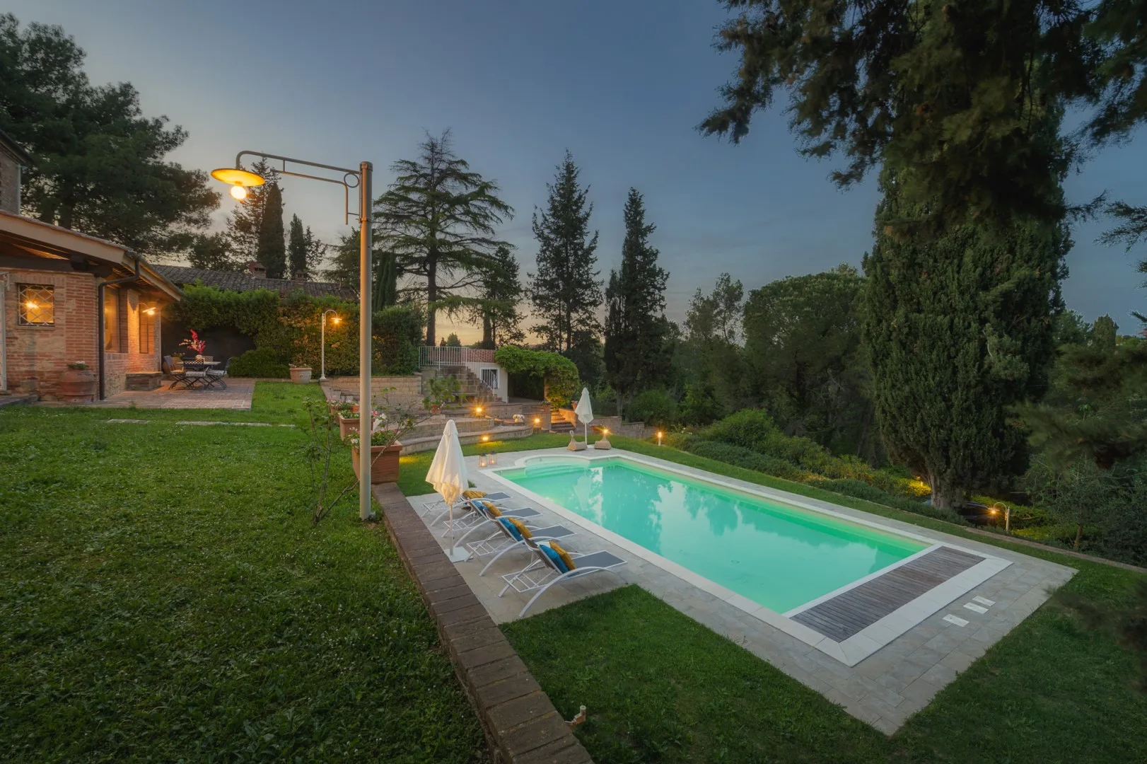 Una piscina al aire libre junto a un césped, con árboles y un patio iluminado.