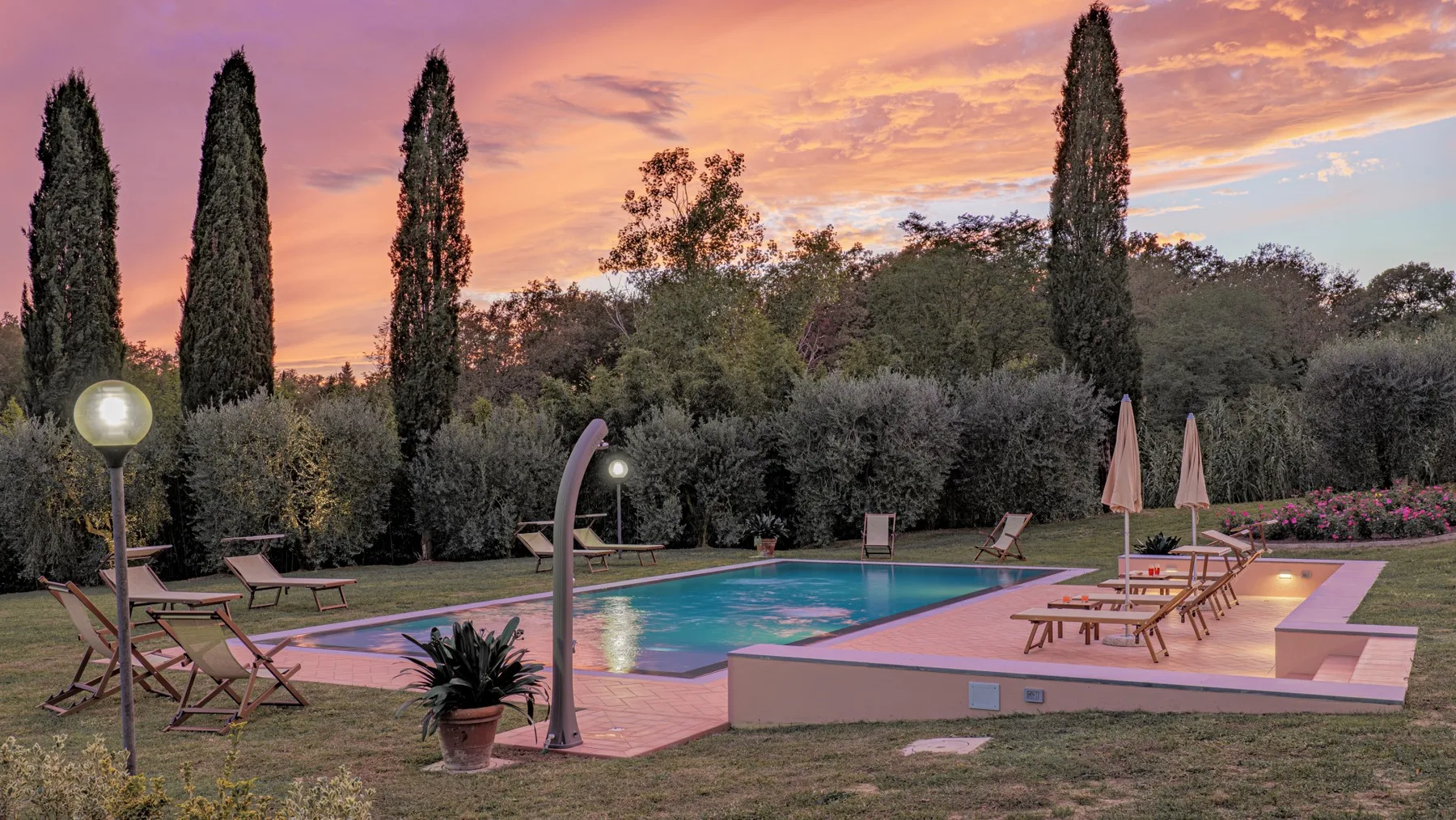 An outdoor swimming pool sits beside a patio with loungers beneath a colorful sunset sky.