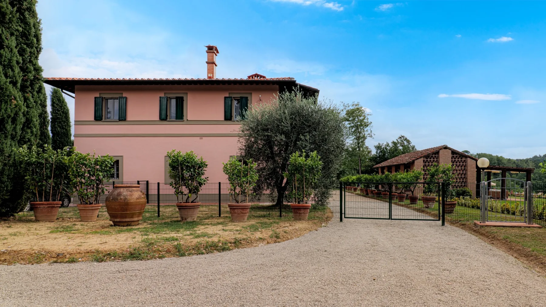 A pink two-story house stands behind a gated gravel driveway, with trees and shrubs.