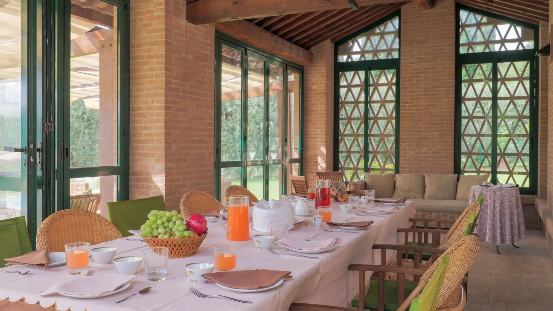 A dining room with a long set table, wicker seating, and large windows.