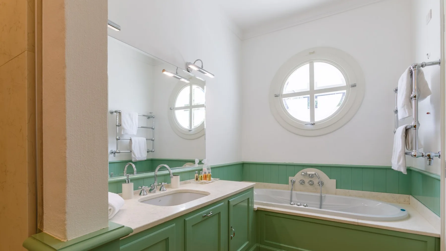 Un baño luminoso tiene muebles verdes, una bañera y una ventana ovalada.