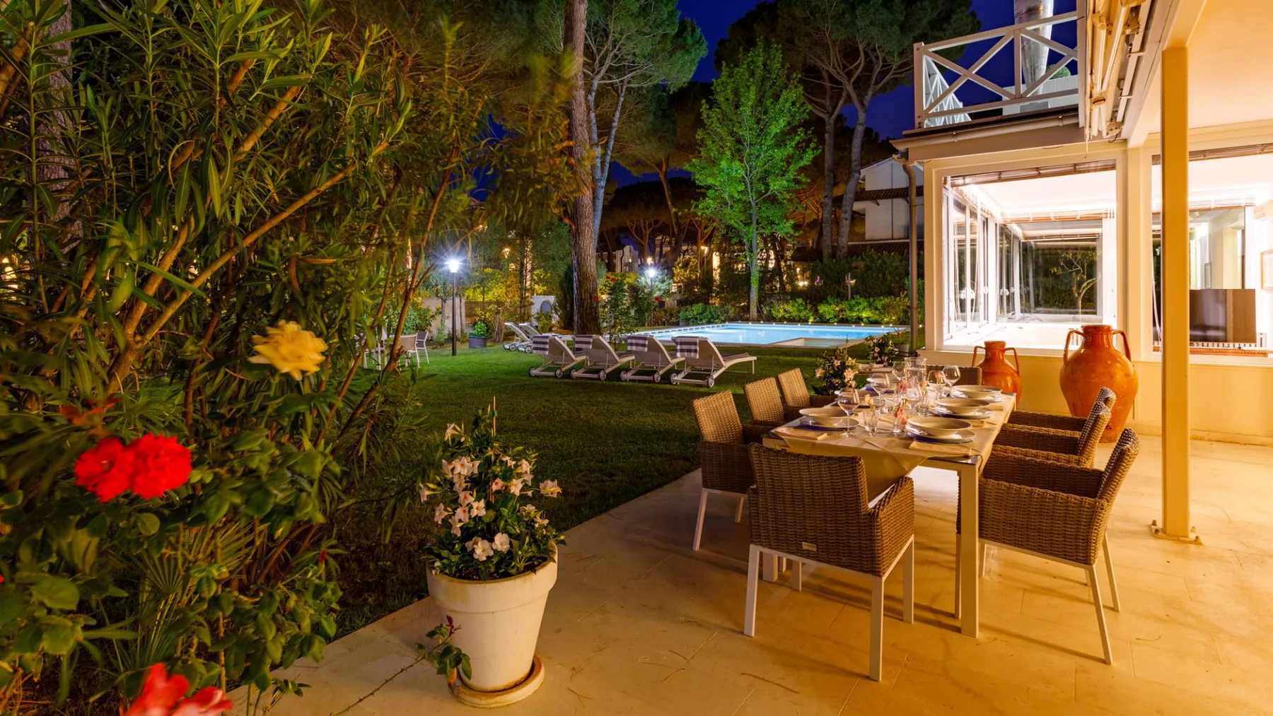 Una terraza comedor exterior da a un jardín y una piscina iluminada.