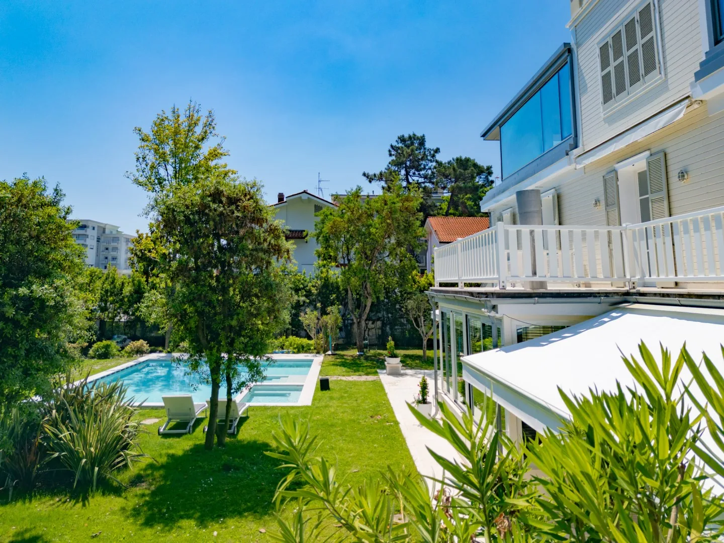 Zona de piscina exterior ajardinada junto a un edificio blanco, con árboles y césped.