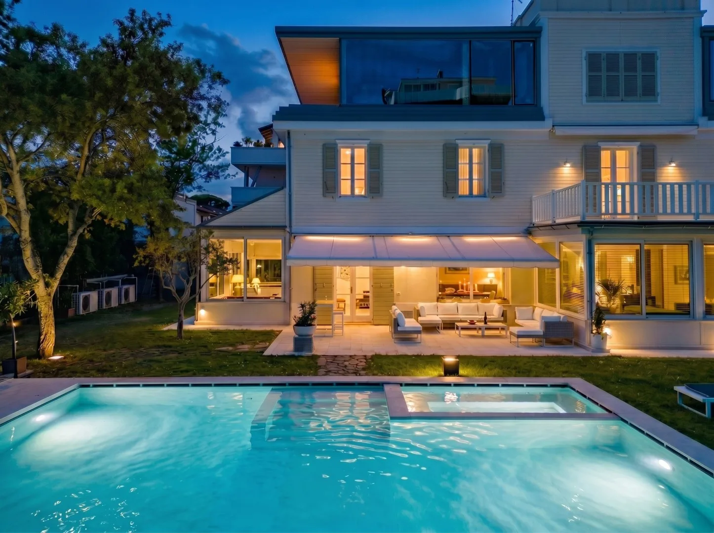 Una casa moderna iluminada al anochecer se alza detrás de una piscina.