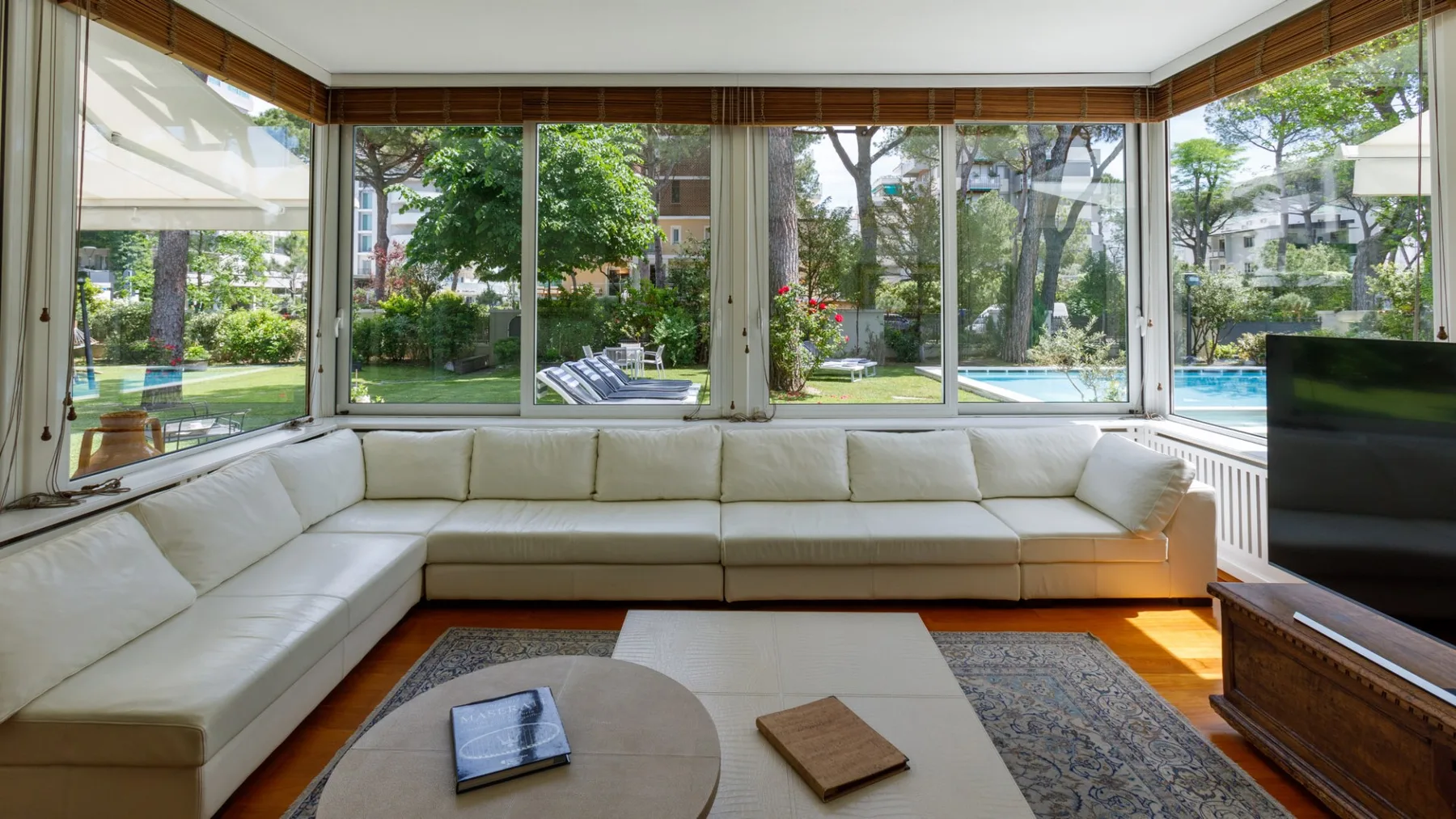Una sala luminosa tiene ventanas panorámicas con vistas a un jardín y una piscina.