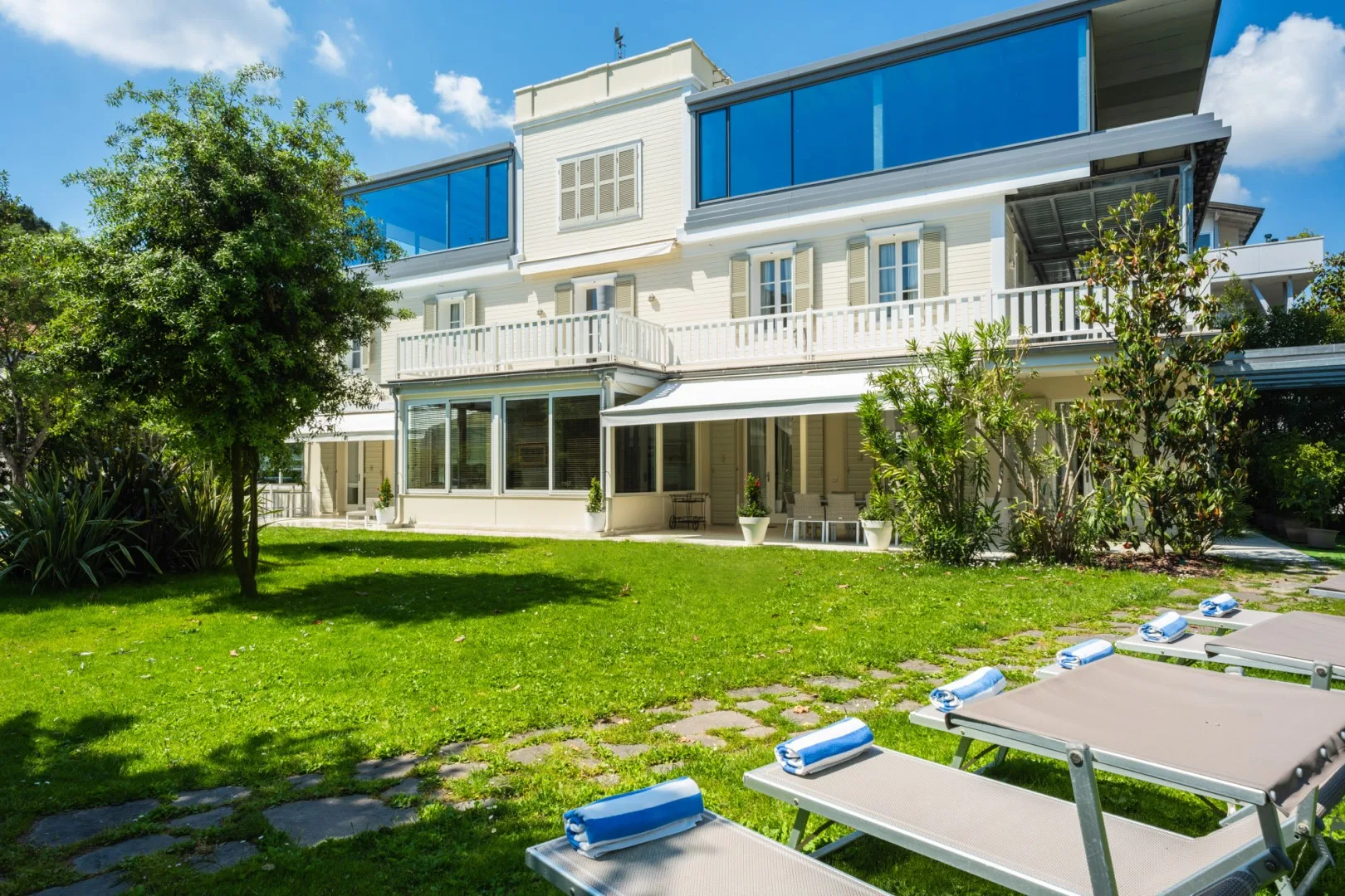 Una casa moderna grande se alza detrás de un césped verde y tumbonas de exterior.