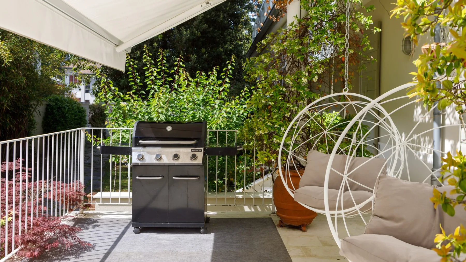 Un patio exterior cubierto tiene una parrilla y asientos acolchados entre plantas frondosas.