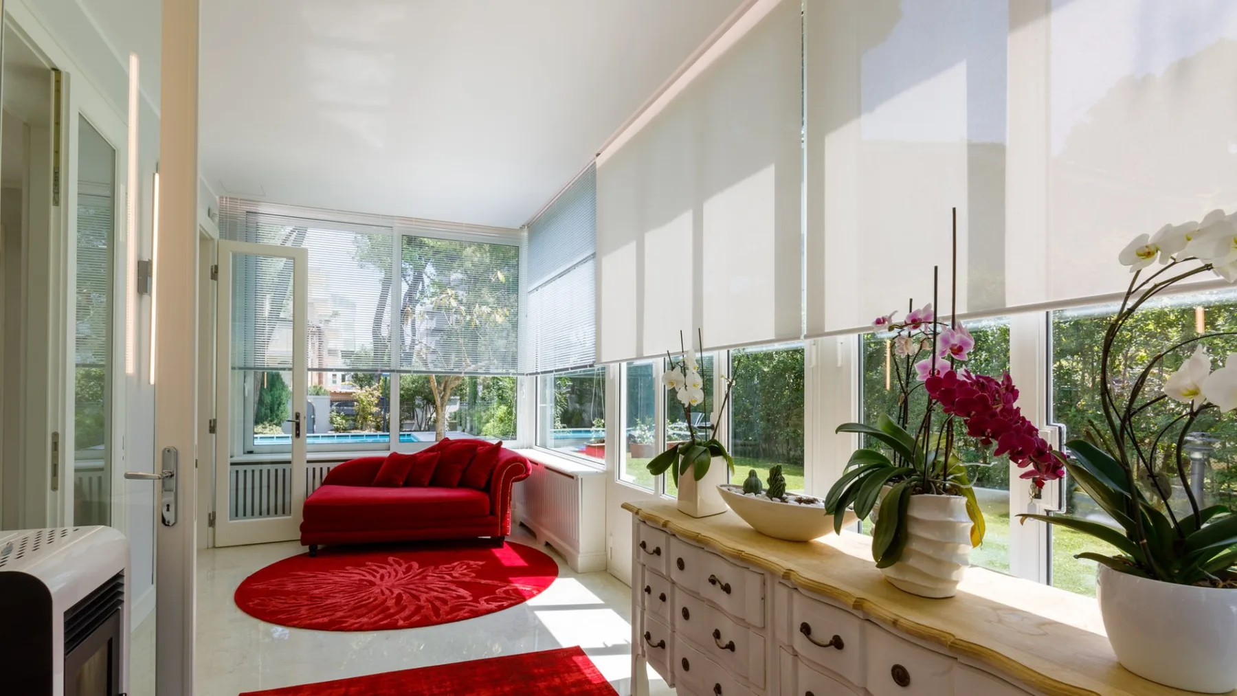 Un solárium luminoso con un sofá rojo, plantas y grandes ventanales.
