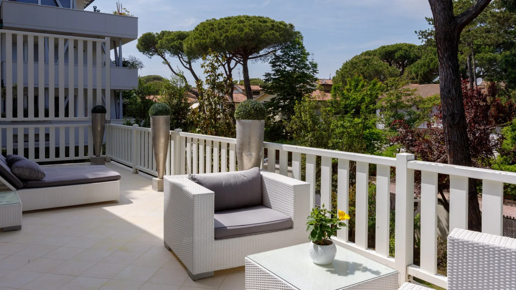 Una terraza exterior luminosa tiene asientos de mimbre y flores en macetas junto a una barandilla blanca.