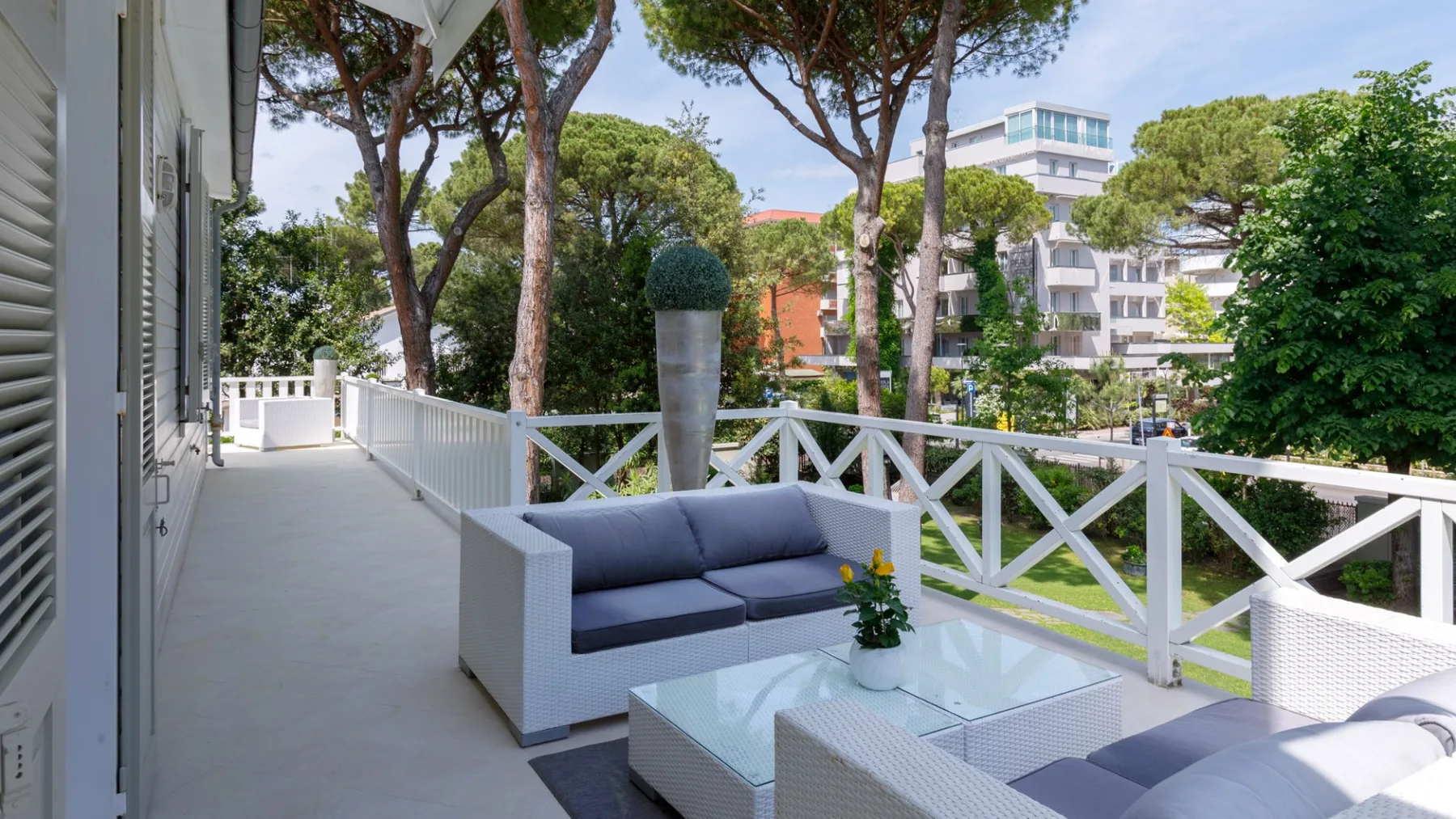 Un patio exterior tiene asientos acolchados junto a una barandilla blanca y árboles frondosos.