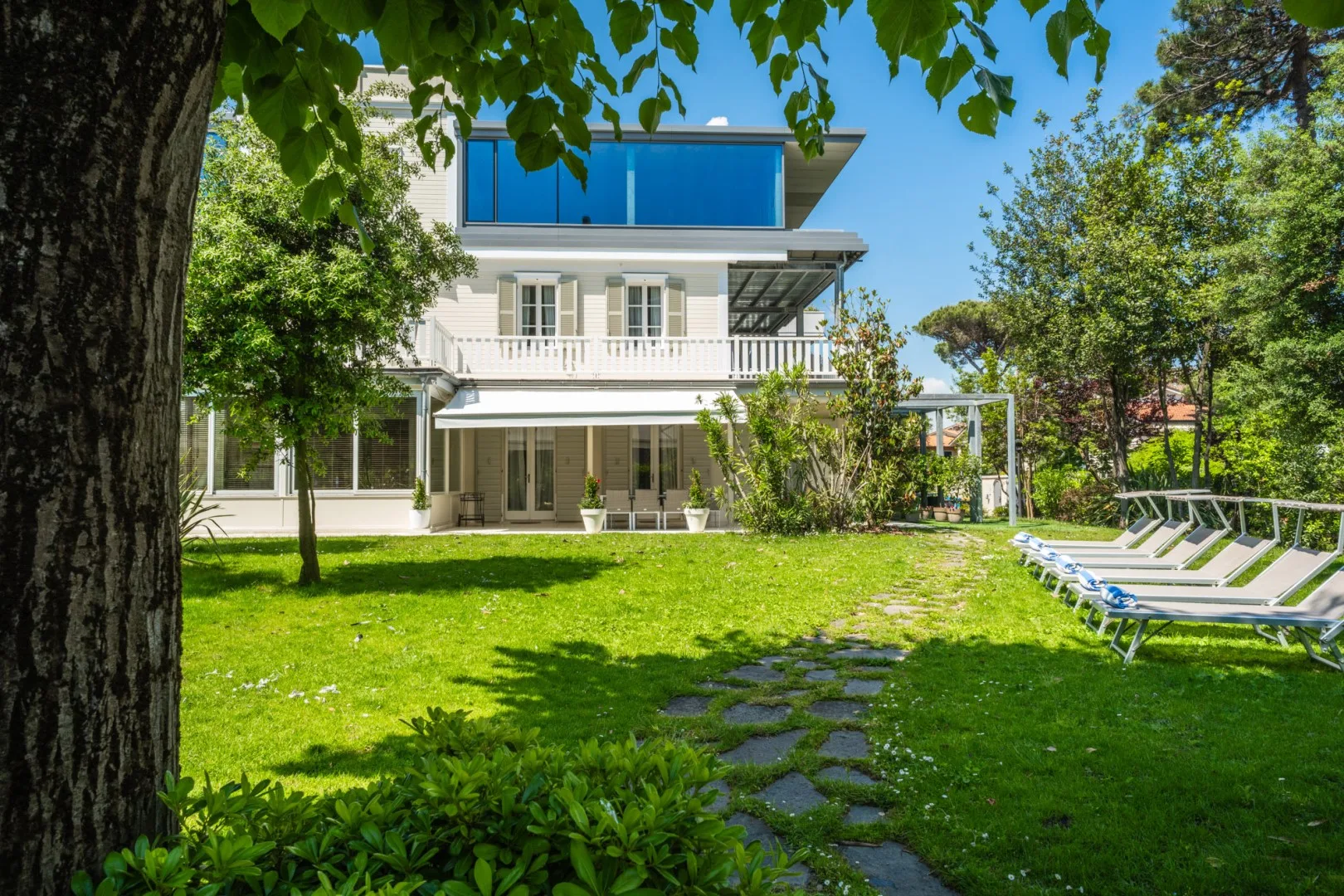 Una casa moderna se abre a un césped verde con un camino de piedra y tumbonas al aire libre.