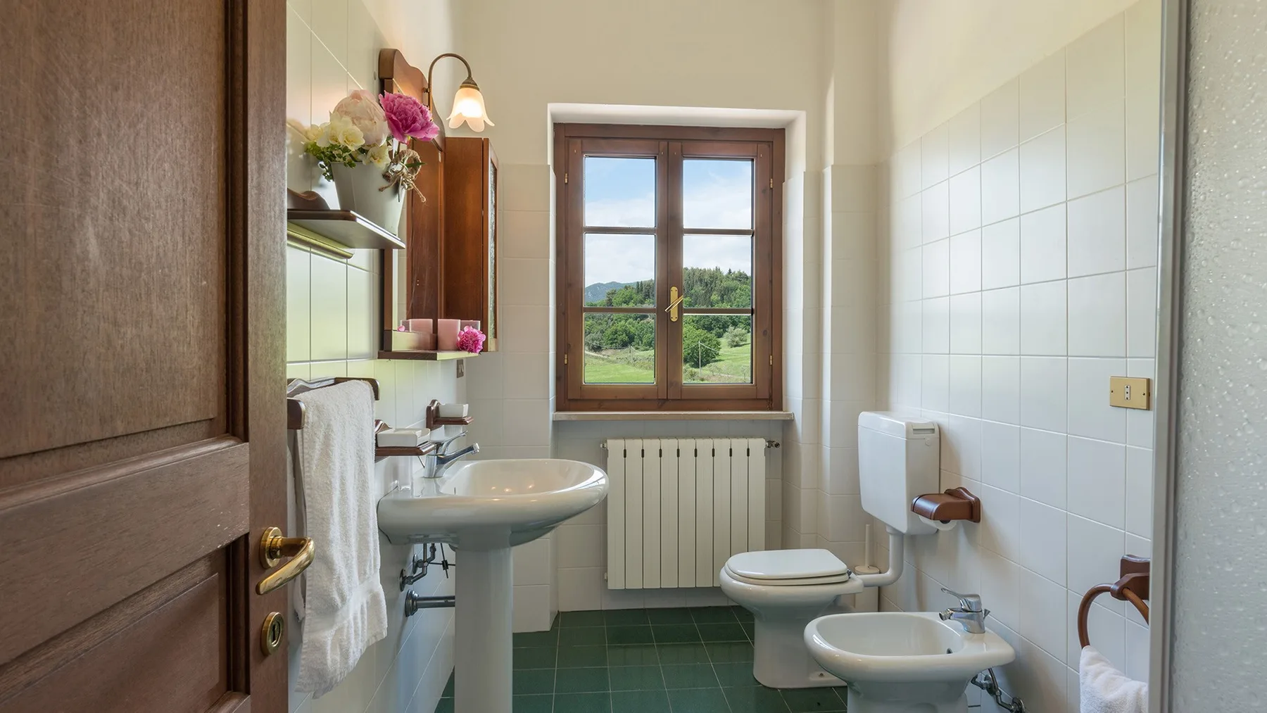 Un baño alicatado con lavabo de pedestal, inodoro, bidé y una ventana con vistas al verde.