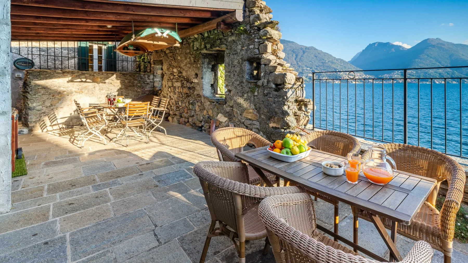 A stone terrace has outdoor seating and a table overlooking a lake and mountains.