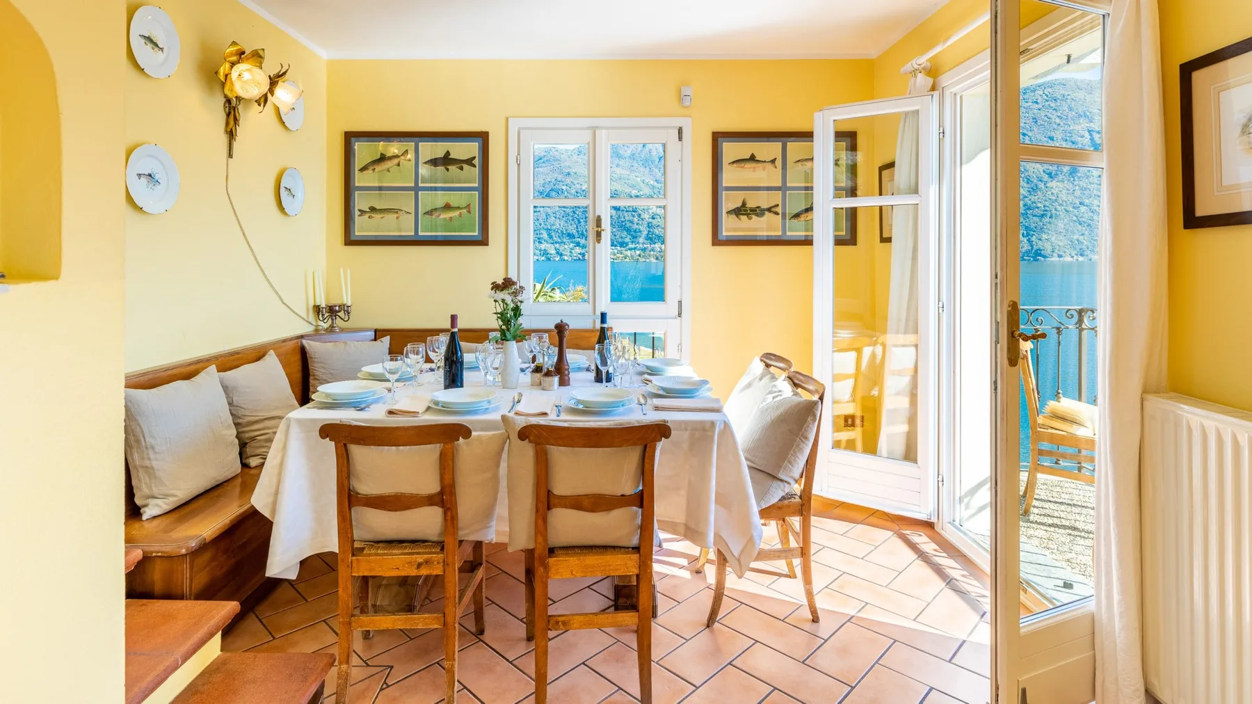 A bright dining room has a set table beside open doors overlooking the water.