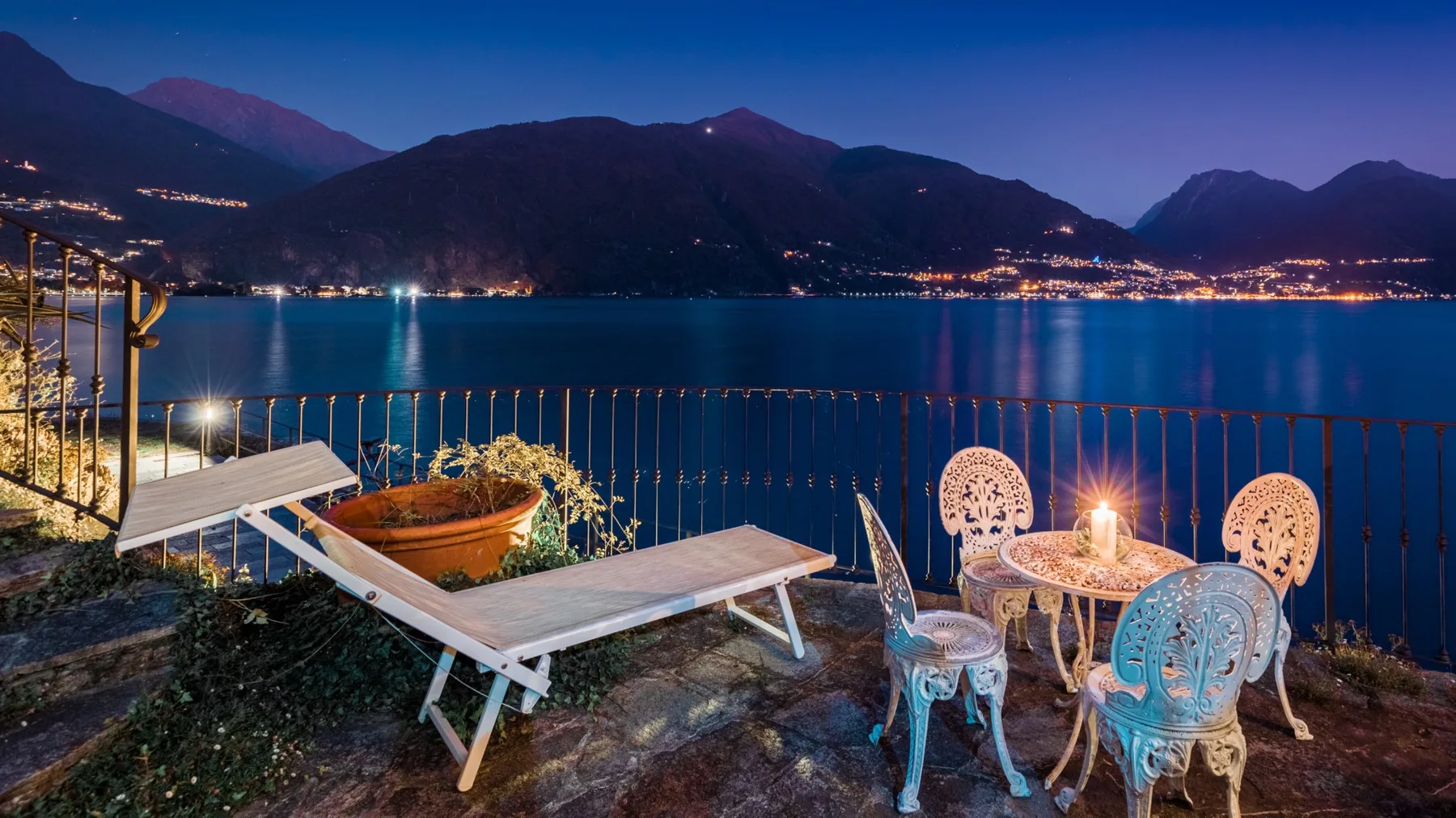 A lakeside terrace at dusk has outdoor seating overlooking the water and illuminated mountains.