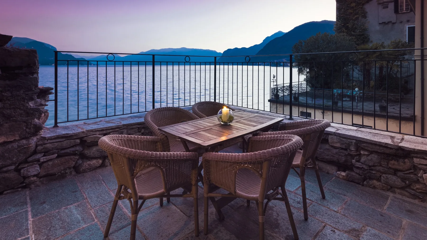 A stone terrace overlooks the water, with wicker seating around a table.