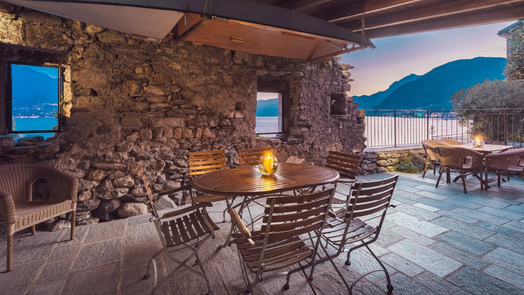 A stone-walled outdoor dining area opens onto a terrace overlooking the water and mountains.