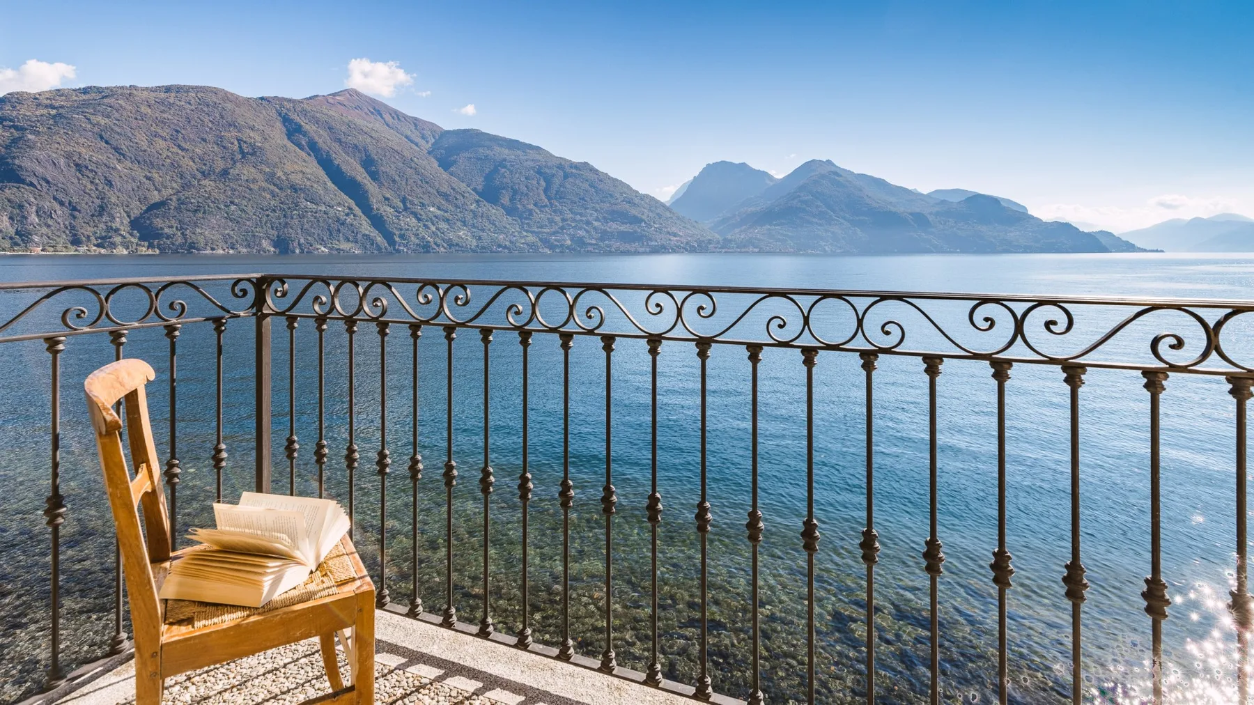 A lakeside balcony overlooks blue water and mountains, with a chair beside the railing.