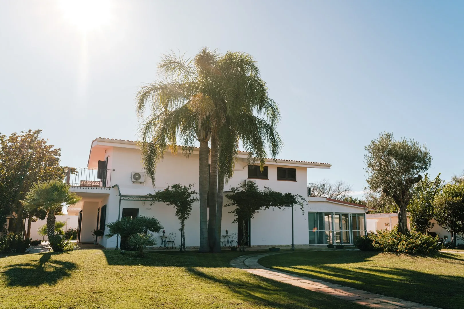 A white house stands behind a green lawn, with palm trees and a walkway.