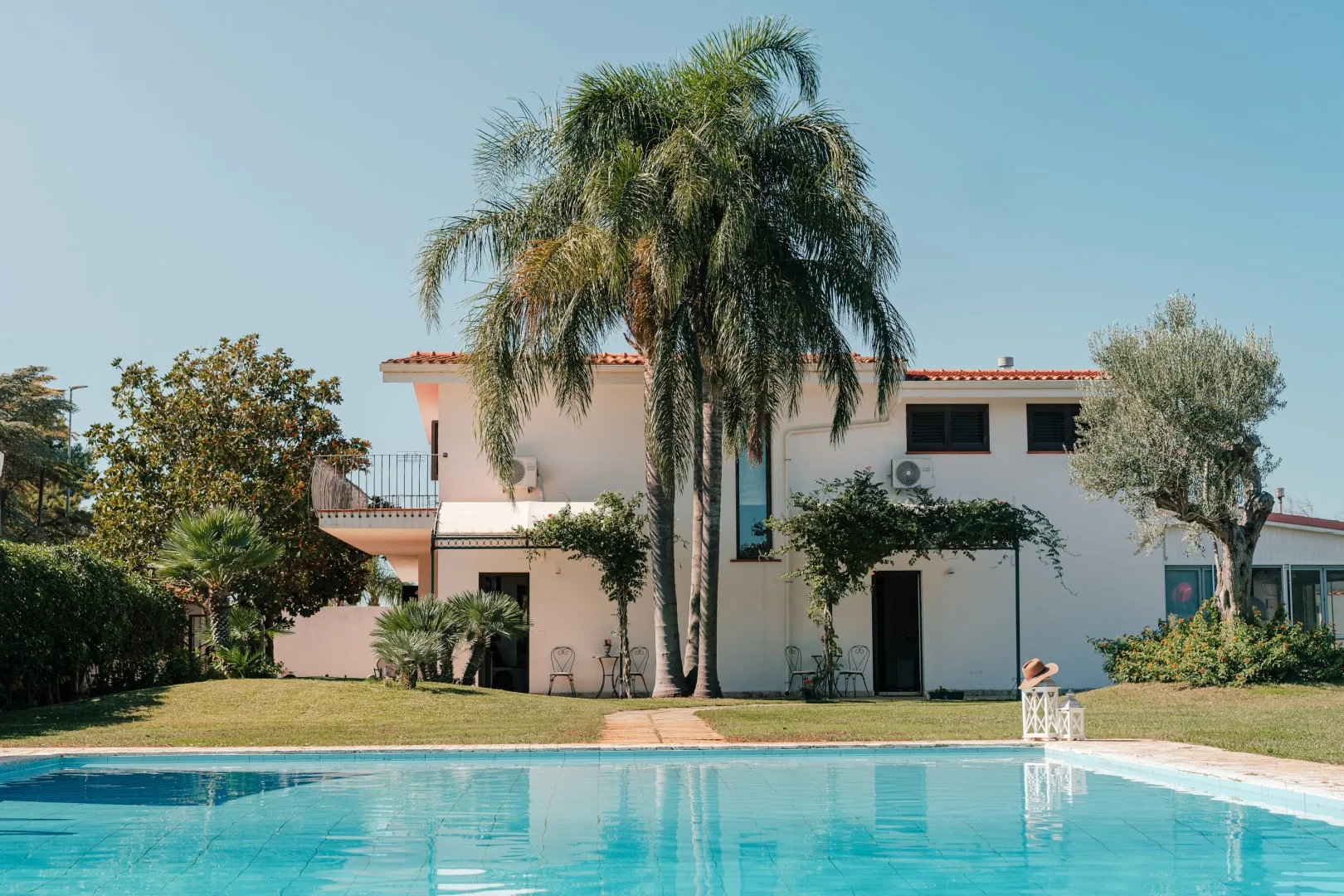 A swimming pool sits in front of a white house with palm trees.