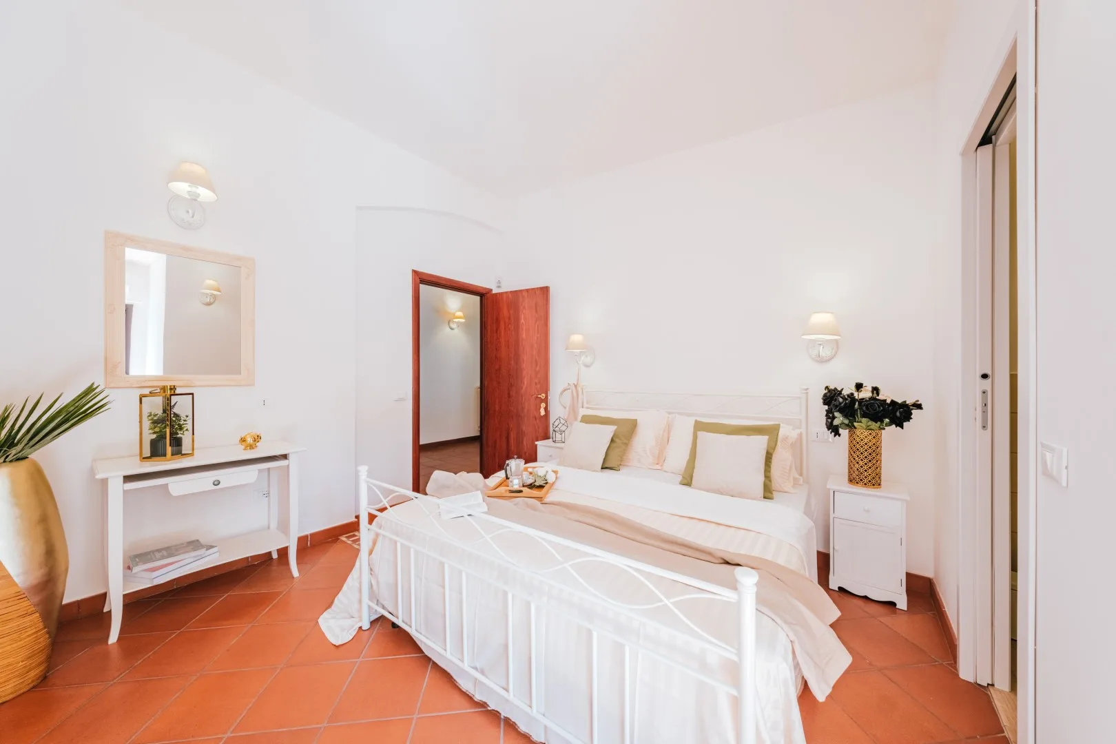 A bright bedroom with a white bed, bedside tables, and terracotta-colored floor tiles.