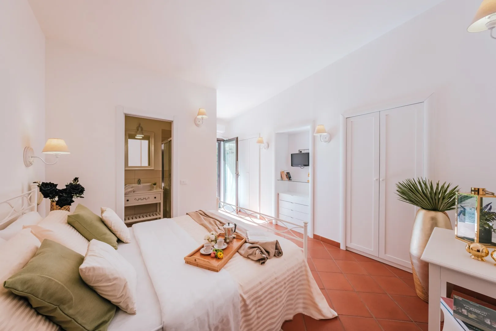 A bright bedroom with a bed, bedside lamps, and a bathroom vanity visible through the doorway.