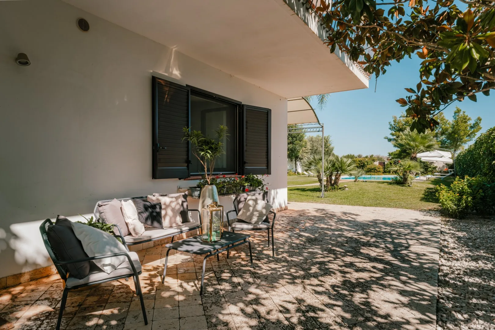 A shaded patio with outdoor seating beside a house and a leafy garden.