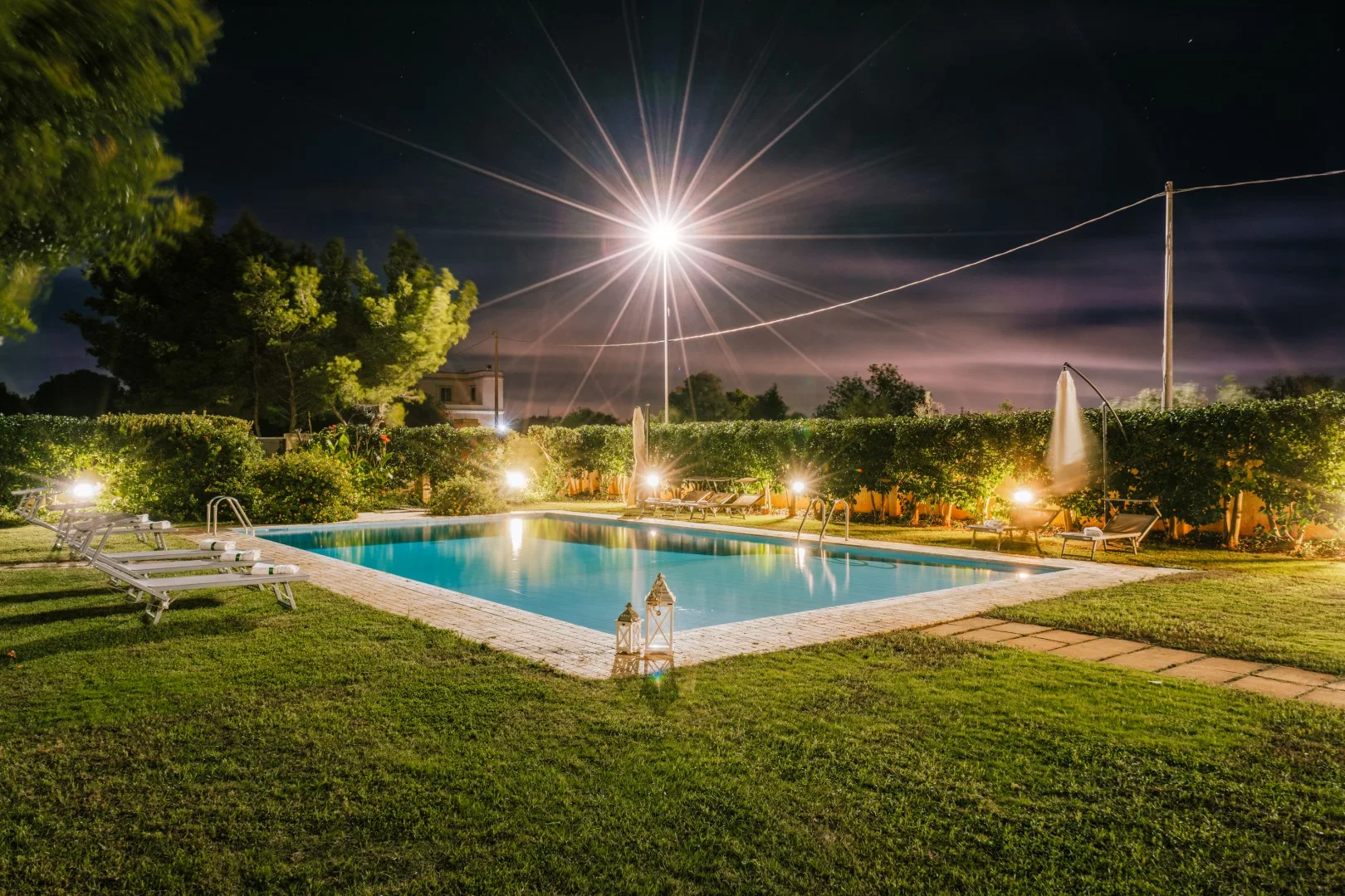 A swimming pool at night is surrounded by lawn, lounge chairs, and trees.