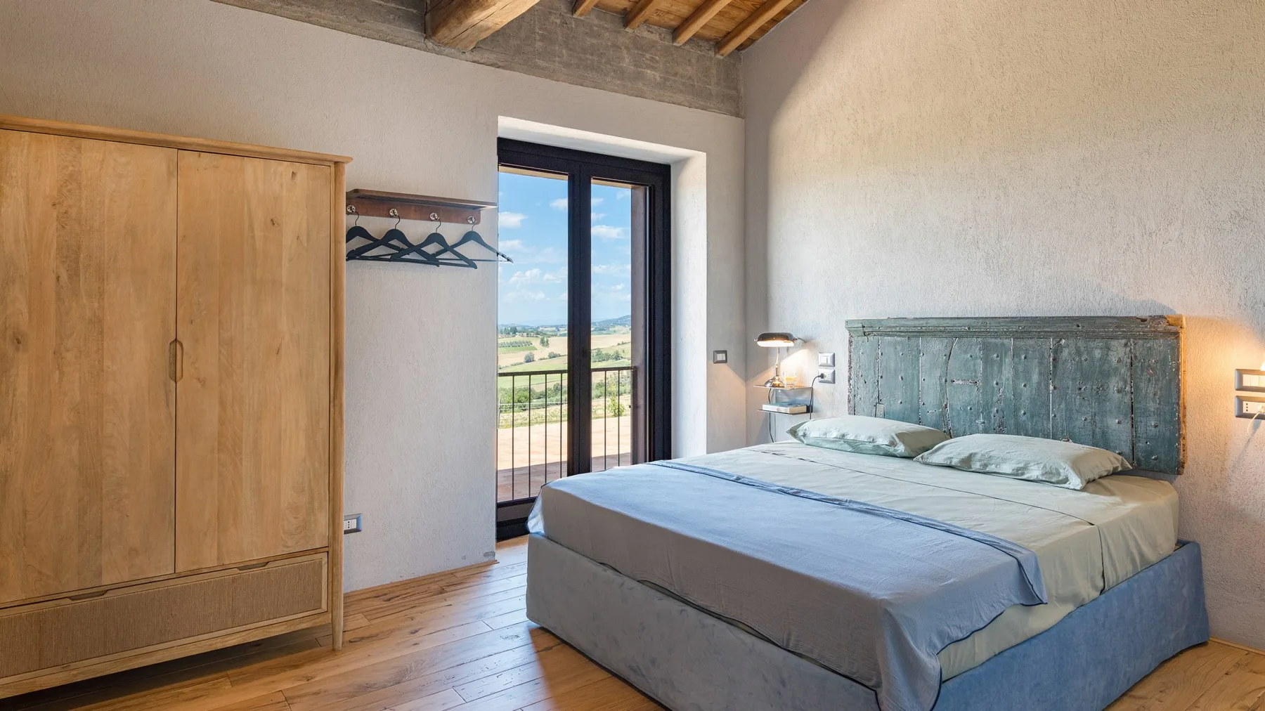 A bedroom with a double bed, a wooden wardrobe, and a balcony door beneath exposed timber beams.