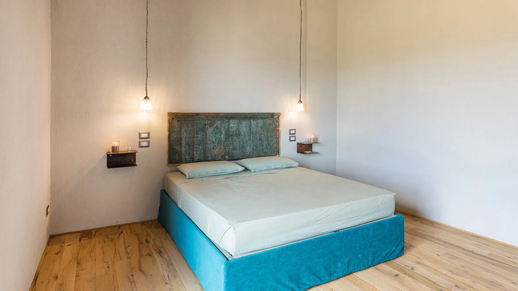 A bedroom with a low bed, turquoise base, pale bedding, and two hanging pendant lights.