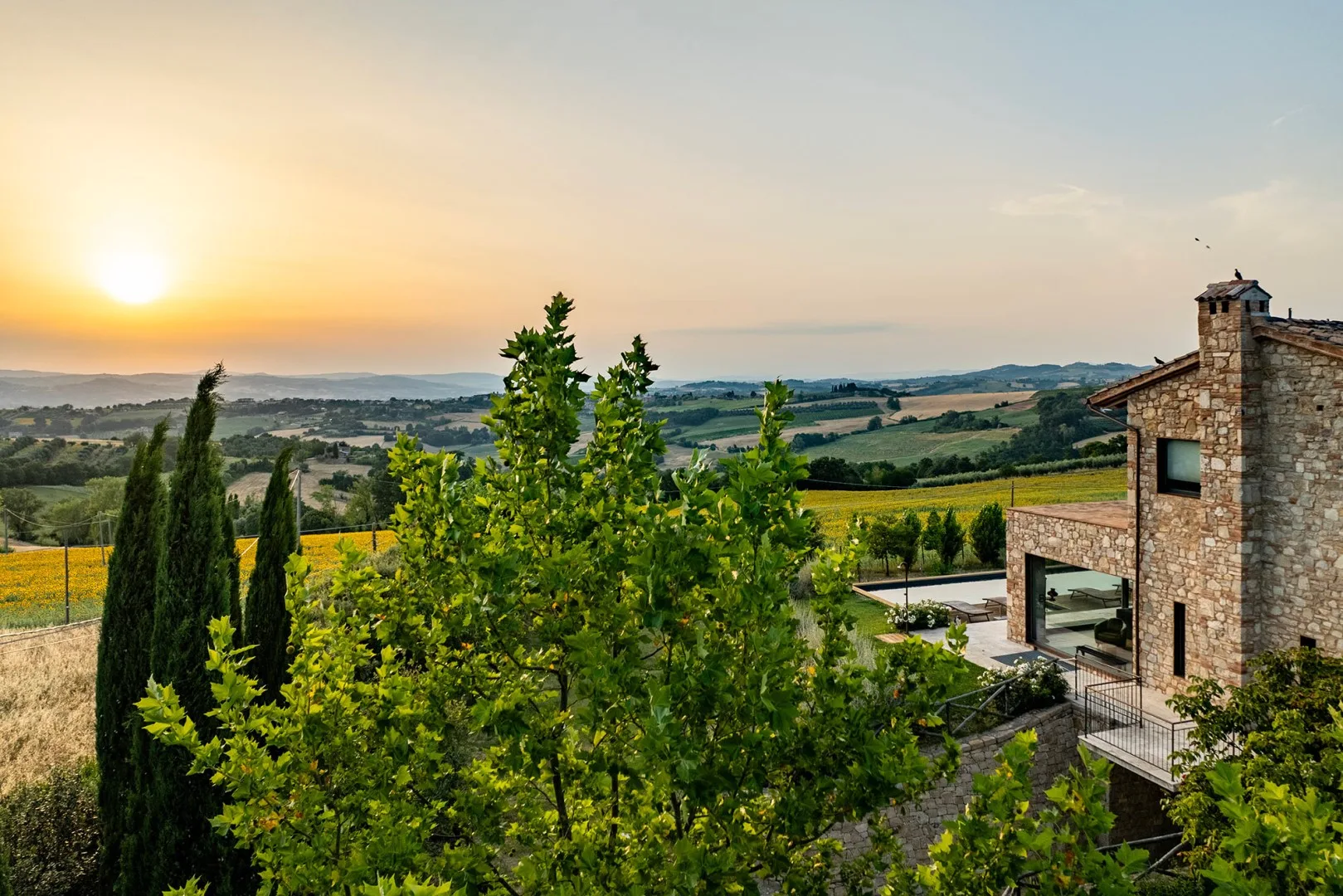 A stone house overlooks rolling fields and hills at sunset, with trees and a garden in the foreground.