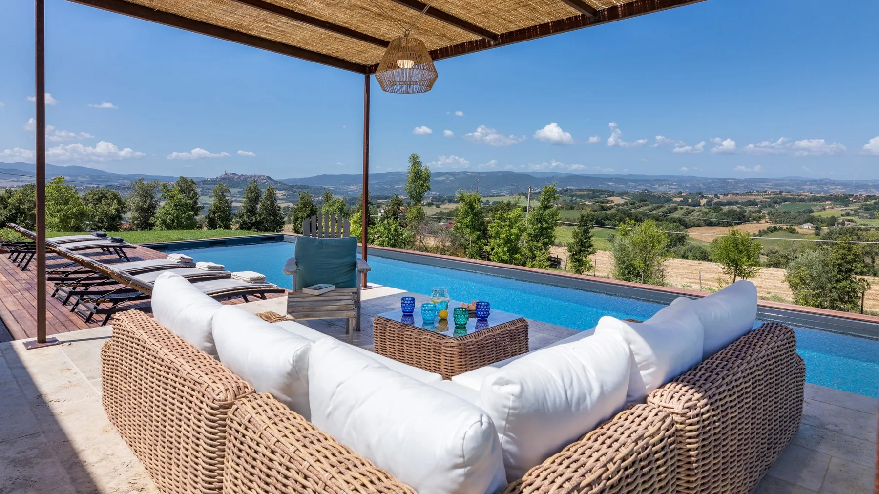 A covered outdoor lounge with wicker sofas and a coffee table overlooks a swimming pool and rolling hills.